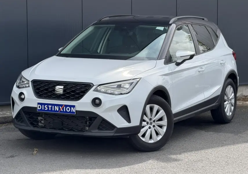 Vue 3/4 avant droite d'un SEAT Arona blanc alpin avec toit noir et jantes alliage 16 pouces, sur fond urbain.
