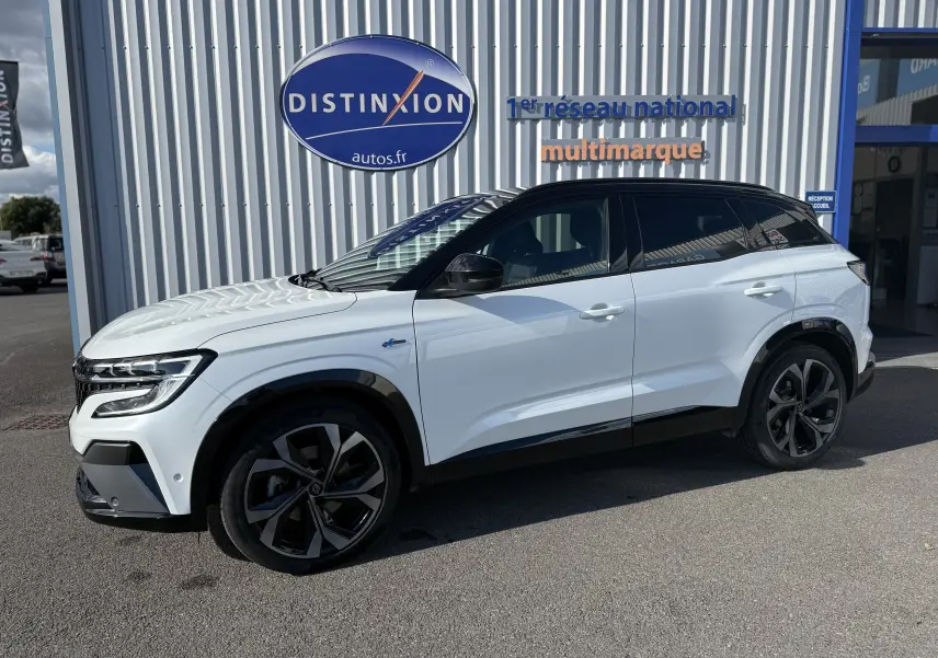 Vue de profil côté gauche du Renault Austral blanc nacré avec toit noir, jantes noires et détails Alpine devant un bâtiment.