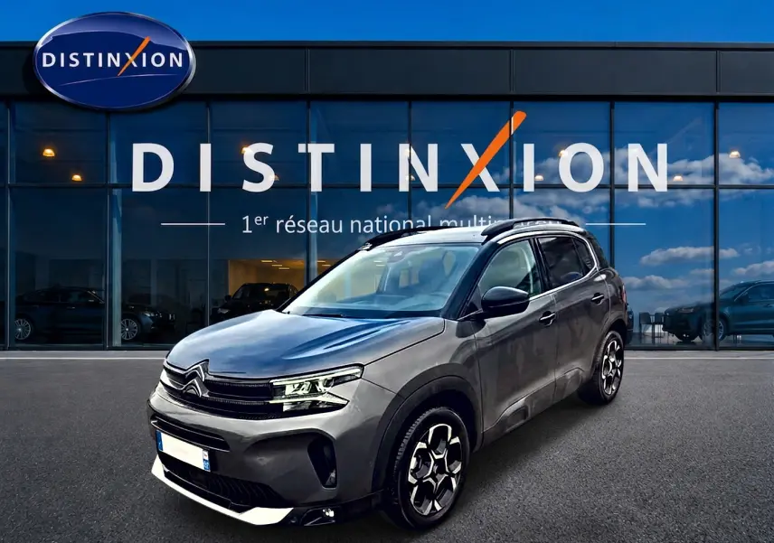 SUV Citroën C5 Aircross gris vu en 3/4 avant droit, avec jantes alu et barres de toit visibles devant un showroom.