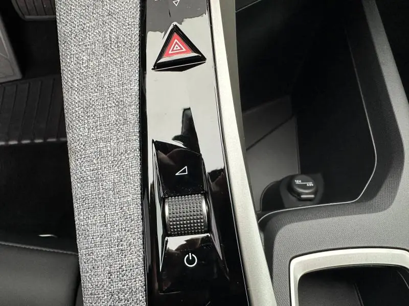 Gros plan sur la console centrale noire brillante avec molette et bouton triangle rouge du Peugeot 3008 Hybrid 2025.