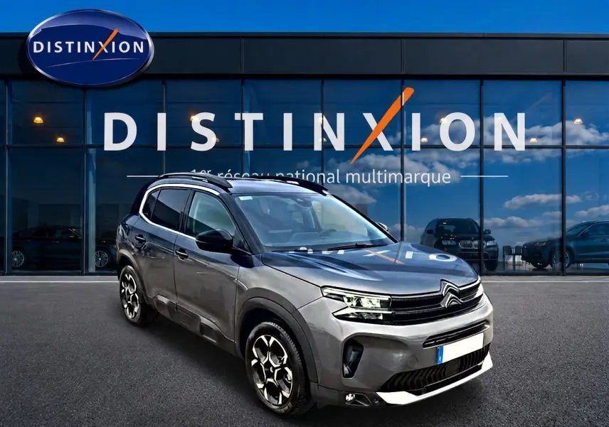 Citroën C5 Aircross gris vue 3/4 avant droit, avec jantes alu et barres de toit visibles devant vitrine Distinxion.