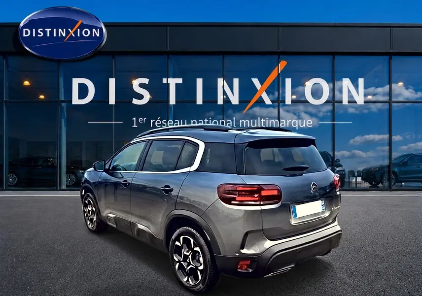 Citroën C5 Aircross gris en 3/4 arrière droit, avec jantes alu et barres de toit visibles devant un showroom Distinxion.