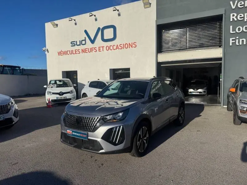 Peugeot 2008 Hybrid gris en 3/4 avant droit, avec barres de toit et jantes alliage 17 pouces, devant un garage sud VO.