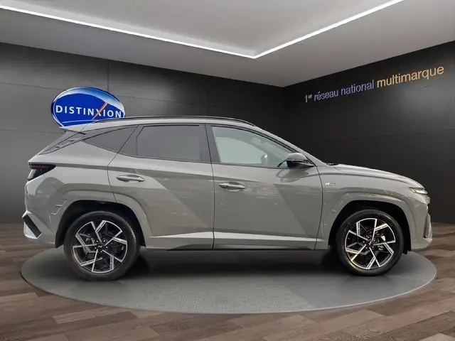 Vue latérale droite d’un Hyundai Tucson 1.6 Hybrid gris foncé, avec jantes alliage et hayon motorisé visible en arrière-plan.