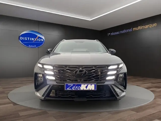 Vue frontale d’un Hyundai Tucson gris foncé 2026 avec feux LED allumés et plaque Zero KM dans un showroom.