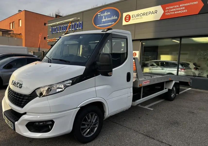 IVECO Daily blanc en 3/4 avant droit, plateau arrière visible, stationné devant un garage avec enseignes.