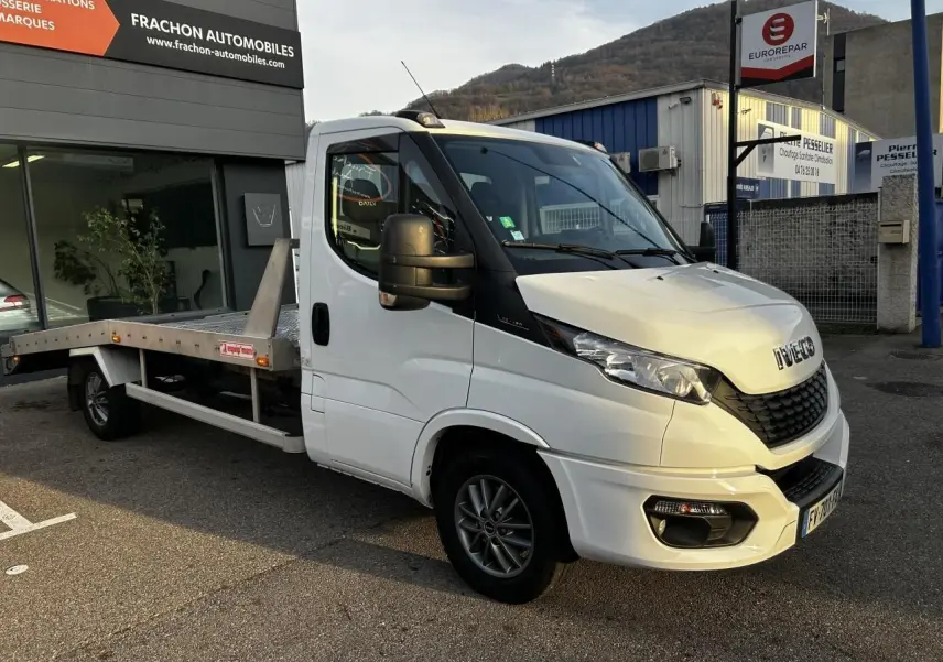 IVECO Daily blanc en 3/4 avant droit, plateau arrière métallique visible, stationné devant un garage automobile.