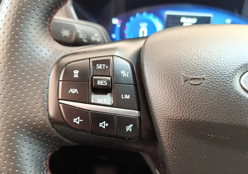 Gros plan sur les commandes gauche du volant en cuir noir perforé du Ford Kuga hybride 2023, avec tableau de bord digital bleu en arrière-plan.