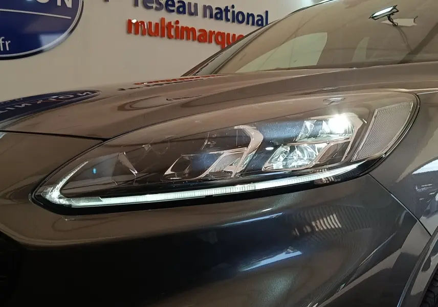 Gros plan sur le phare avant droit allumé du Ford Kuga gris foncé, mettant en valeur les LED et la carrosserie brillante.