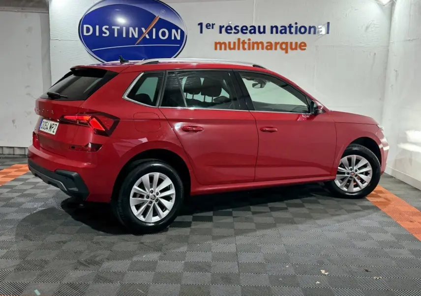 Vue latérale droite d'un Skoda Kamiq rouge 2024 avec jantes alliage et barres de toit noires en intérieur showroom.