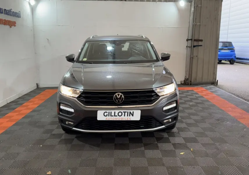 Vue frontale d'un Volkswagen T-Roc gris foncé avec phares allumés et logo VW bien visible sur calandre noire.