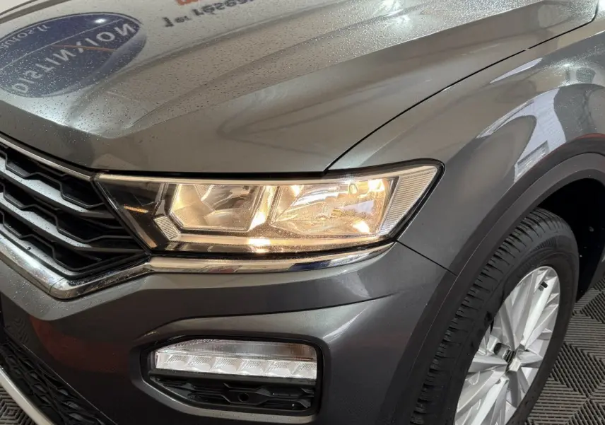 Gros plan sur l'avant droit d'un Volkswagen T-Roc gris foncé avec phares allumés et jante argentée visible.
