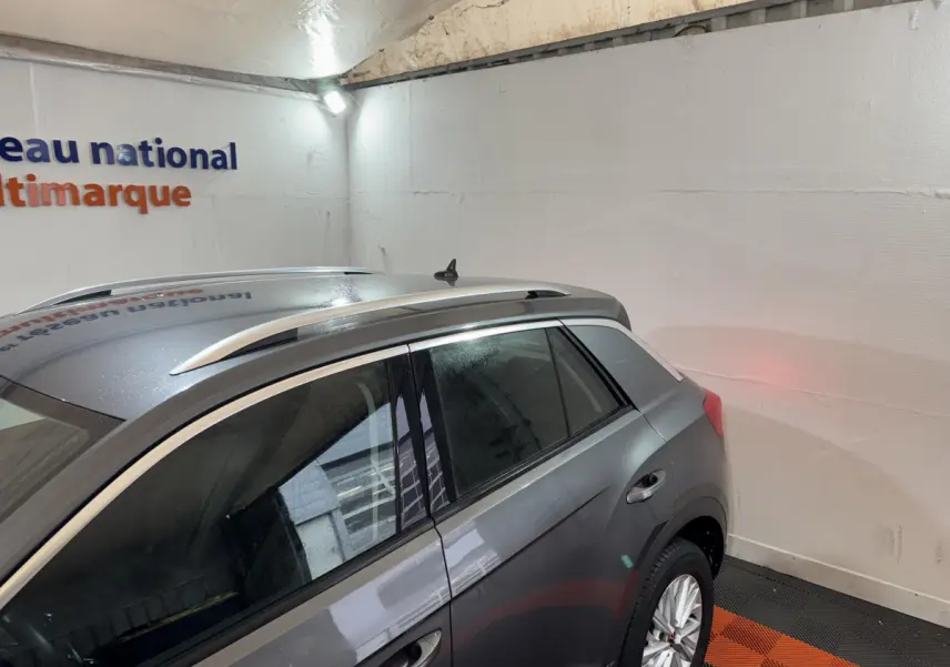 Vue côté gauche arrière du Volkswagen T-Roc gris foncé avec barres de toit argentées dans un garage.