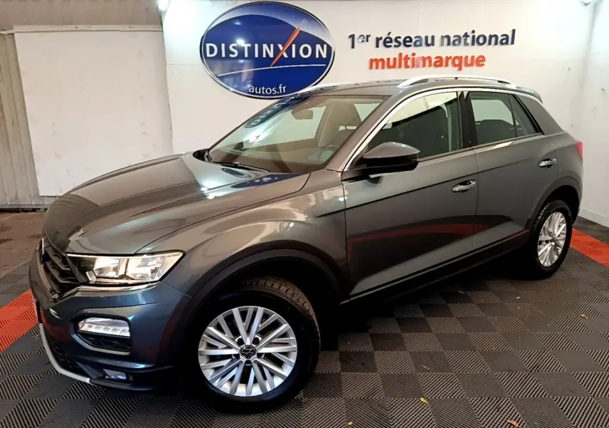 Volkswagen T-Roc gris foncé vue 3/4 avant droit, avec jantes alliage et barres de toit argentées.
