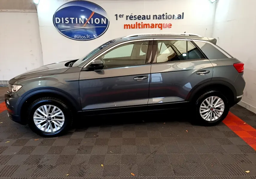 Profil côté gauche d'un Volkswagen T-Roc gris foncé 2021 avec jantes alliage et rails de toit blancs dans un showroom.