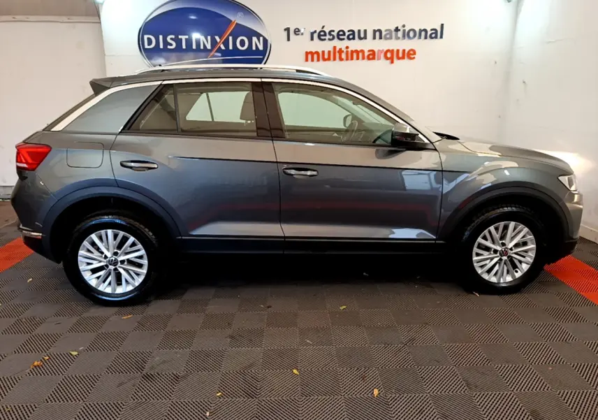 Profil côté gauche d’un Volkswagen T-Roc gris foncé 2021 dans un showroom avec sol à damier et logo Distinxion en arrière-plan