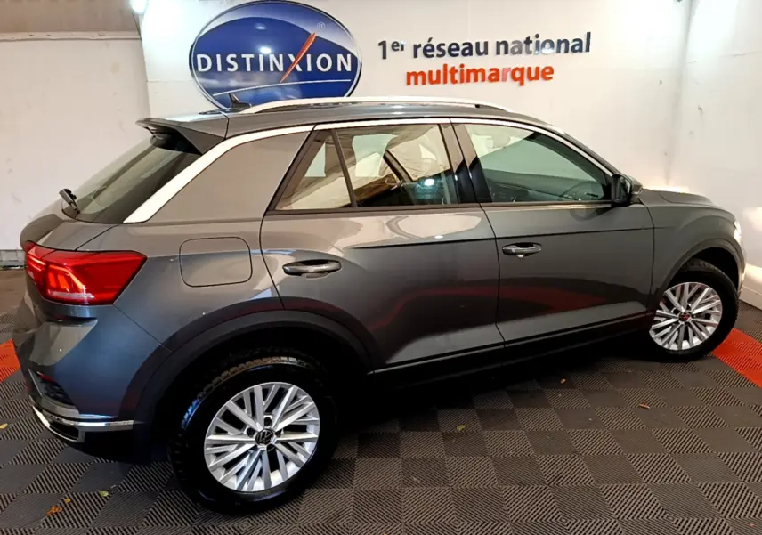 Vue de profil côté droit d'un Volkswagen T-Roc gris foncé avec jantes alliage et rails de toit blancs dans un showroom.