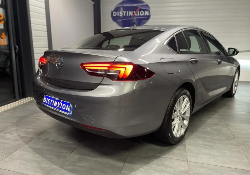 Opel Insignia Grand Sport gris clair vue 3/4 arrière droit avec feux arrière LED allumés en intérieur showroom.