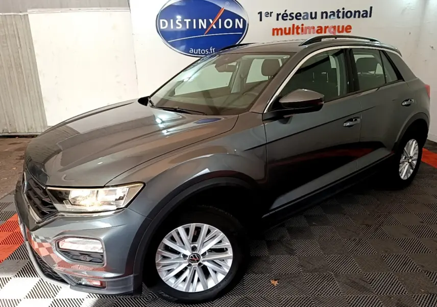 Volkswagen T-Roc gris foncé vue 3/4 avant droit, avec jantes argentées et phares allumés en intérieur showroom.