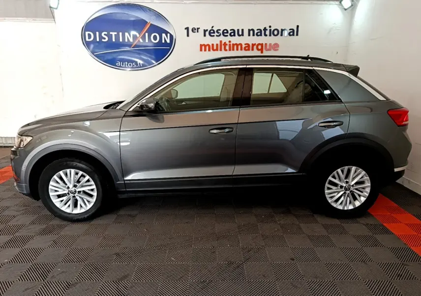 Profil côté gauche d'un Volkswagen T-Roc gris foncé 2021 dans un showroom avec logo Distinxion en arrière-plan.