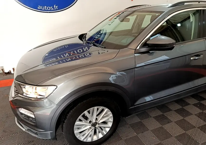 Vue 3/4 avant droit d'un Volkswagen T-Roc gris foncé avec jantes alliage et feux allumés dans un showroom.