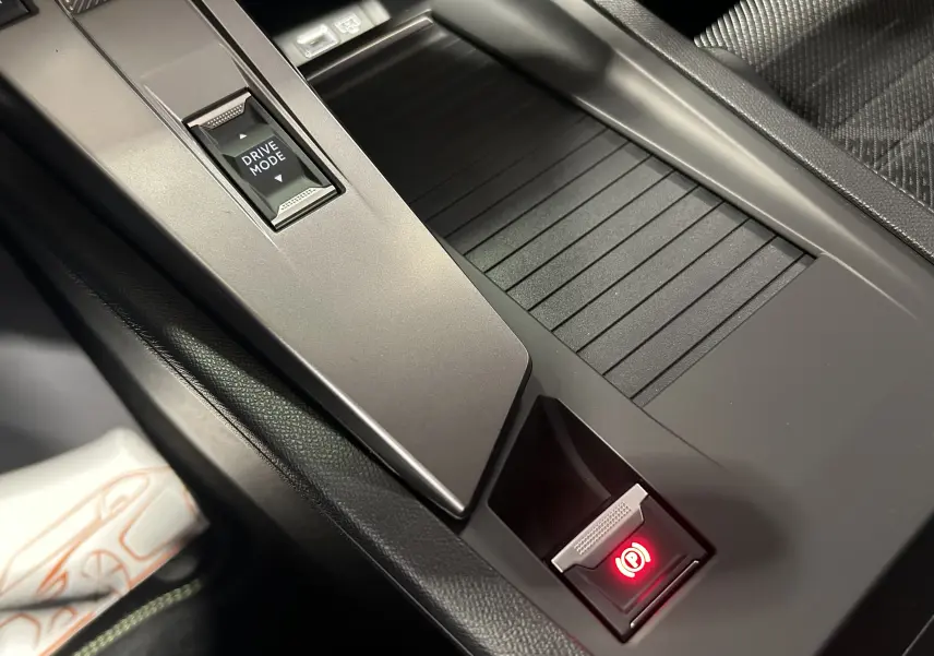 Gros plan sur la console centrale grise de la Peugeot 308 Hybrid 2026, avec bouton frein à main électronique allumé.