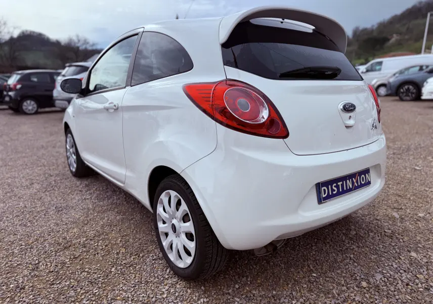 Vue 3/4 arrière droite d'une Ford Ka blanche avec hayon et feux arrière rouges sur un sol gravillonné.