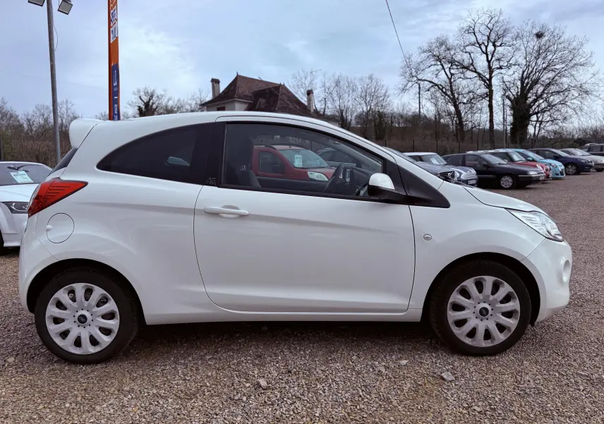 Vue de profil côté gauche d'une Ford Ka blanche 3 portes garée sur un sol gravillonné en extérieur.