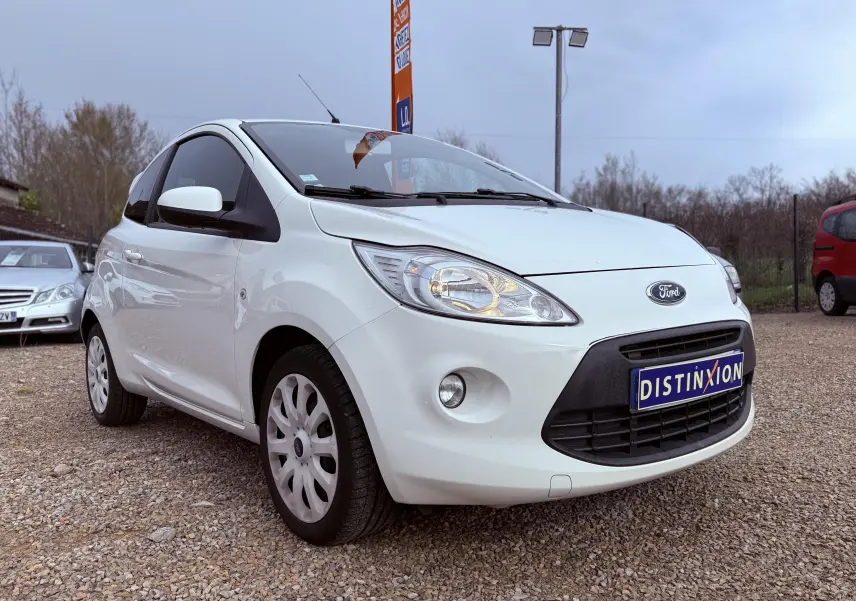 Ford Ka blanc vue 3/4 avant droit sur parking gravillonné avec plaque Distinxion visible.