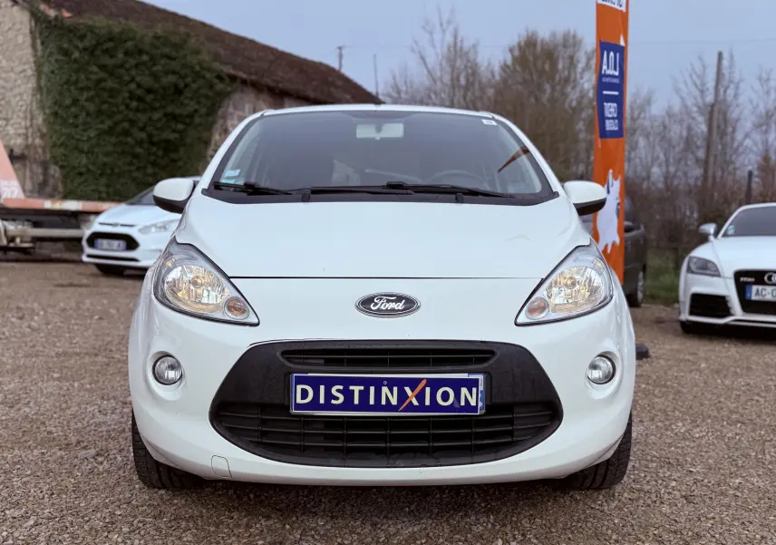 Vue avant d'une Ford Ka blanche avec plaque Distinxion, phare allumé, sur un sol gravillonné en extérieur.