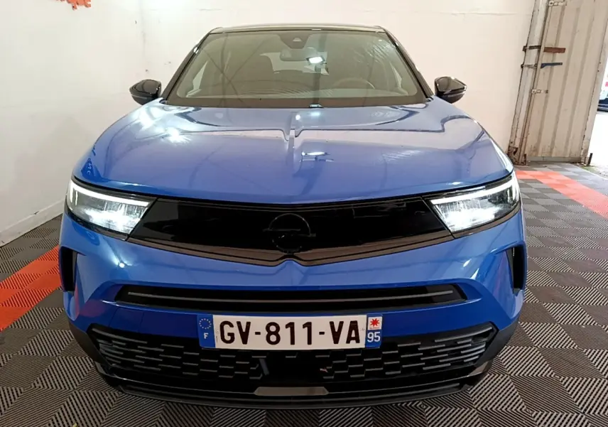 Vue avant d'un Opel Mokka bleu 2024 avec calandre noire et phares LED allumés dans un garage.
