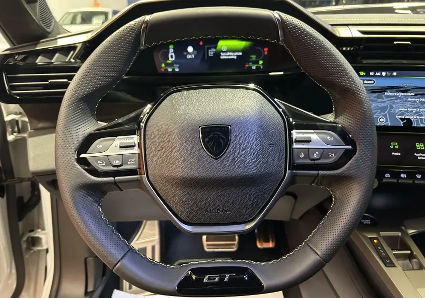 Vue rapprochée du volant cuir perforé noir avec logo Peugeot et badge GT, tableau de bord numérique et écran GPS visibles.