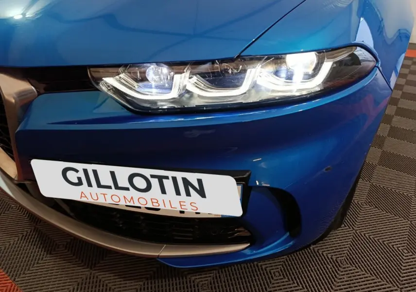 Gros plan sur le phare avant droit allumé d'une Alfa Romeo Tonale bleu 2023 avec calandre et plaque avant visibles.