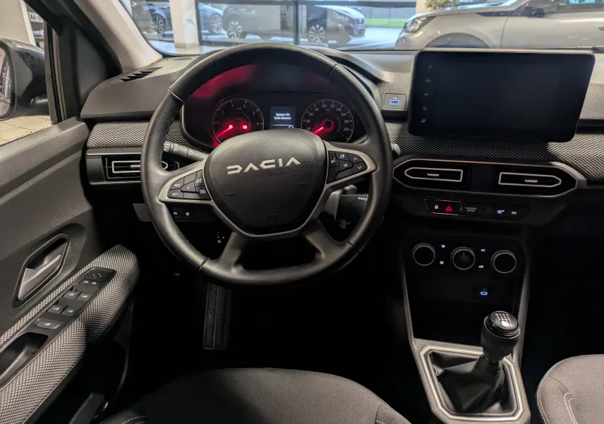 Vue intérieure centrée sur le volant DACIA et tableau de bord avec écran tactile et levier de vitesse manuel.