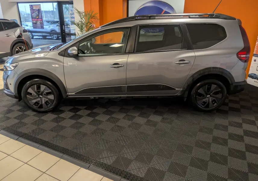 Vue de profil côté gauche d'un Dacia Jogger gris schiste métal, avec jantes noires et intérieur visible en showroom.