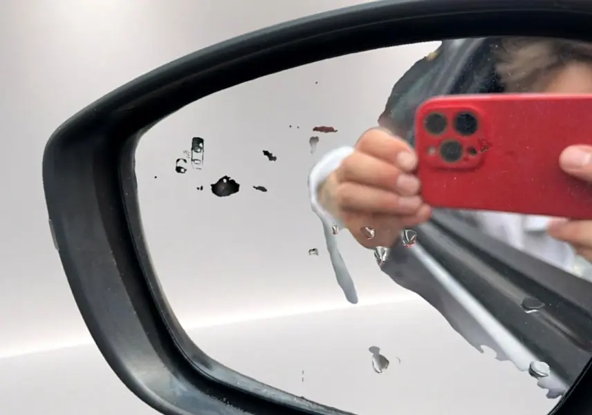 Rétroviseur droit d’un Opel Mokka gris 2025 avec traces d’eau et reflet d’une main tenant un smartphone rouge.
