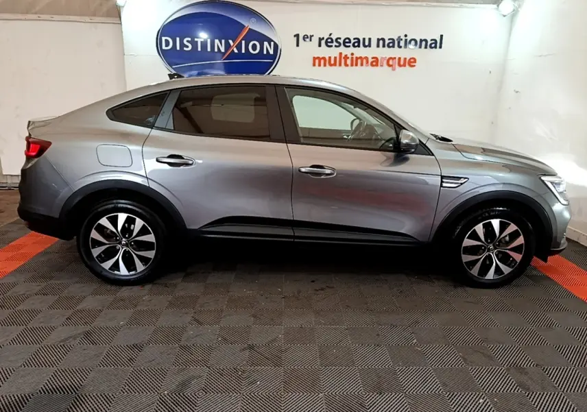 Profil côté gauche d'une Renault Arkana Business E-TECH 145 gris foncé dans un showroom avec logo Distinxion en arrière-plan