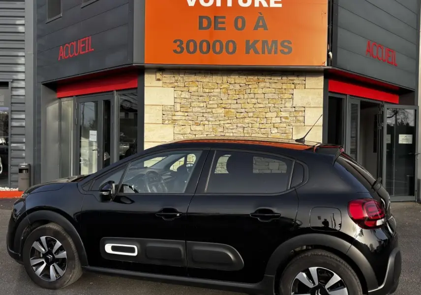 Profil droit d'une Citroën C3 PureTech 110 Shine noire, avec protections latérales et jantes distinctives, devant un bâtiment.