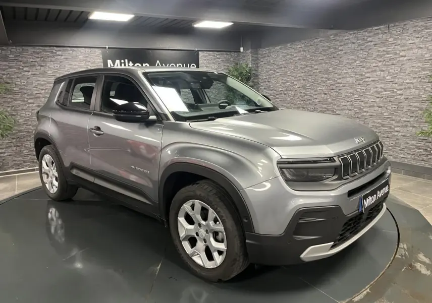 Jeep Avenger gris métallisé en 3/4 avant droit, avec calandre distinctive et coques de rétroviseurs noires brillantes.