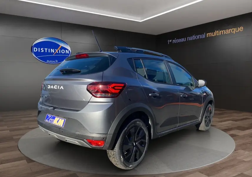 Vue 3/4 arrière droite d'une Dacia Sandero Stepway gris schiste avec barres de toit et feux LED allumés.