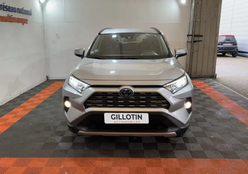 Vue frontale d'un Toyota RAV4 gris clair hybride avec phares et antibrouillards allumés en intérieur showroom.