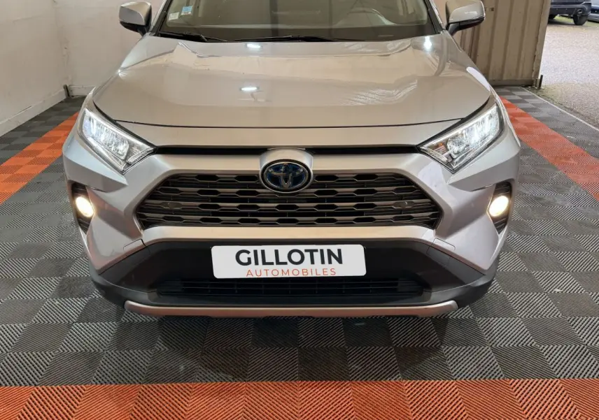 Vue frontale d'un Toyota RAV4 gris clair avec phares et antibrouillards allumés, calandre noire hexagonale.