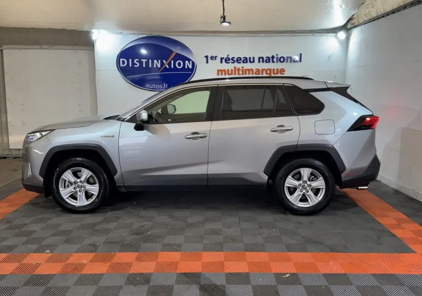 Profil côté gauche d'un Toyota RAV4 hybride gris clair, avec jantes alliage et badges hybrides visibles en intérieur showroom.