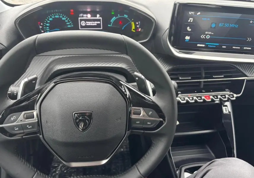 Vue intérieure centrée sur le volant noir d'une Peugeot 208 Hybrid 2025 avec tableau de bord numérique et écran tactile.