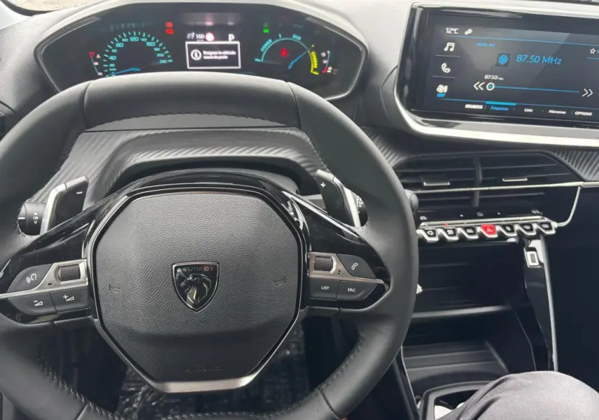 Vue rapprochée du volant et du tableau de bord numérique d'une Peugeot 208 Hybrid gris, avec écran tactile et commandes au volant.