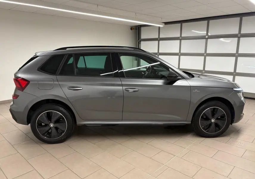 Vue de profil côté gauche d’un Skoda Kamiq 1.5 TSI Monte Carlo gris graphite avec jantes noires distinctives, en intérieur.