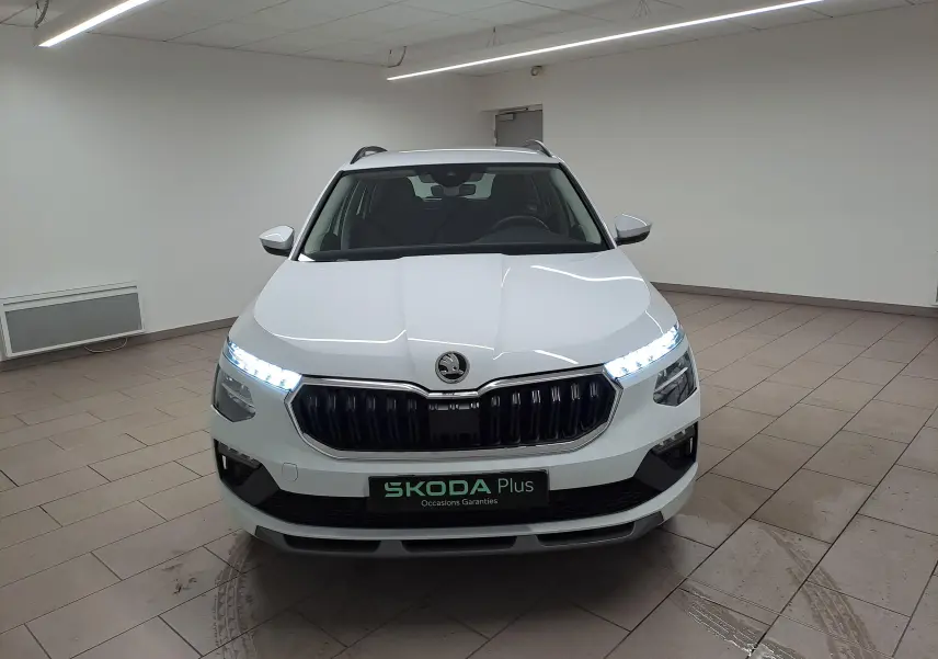 Vue frontale du Skoda Kamiq blanc Lune 2025 avec phares allumés et calandre noire dans un showroom.