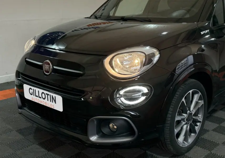 Vue 3/4 avant droit d'une FIAT 500X noire avec phares allumés et logo Sport visible sur l'aile avant.