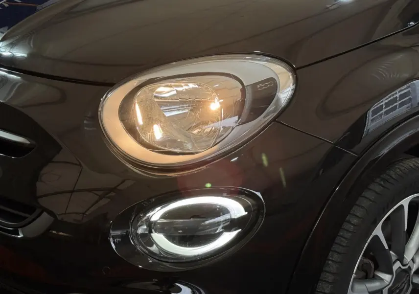 Gros plan sur le phare avant gauche allumé et le feu antibrouillard LED d'une FIAT 500X noire, avec jante visible.