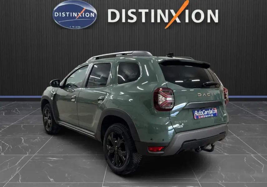 Vue 3/4 arrière droite du Dacia Duster 2024 kaki lichen avec jantes noires et barres de toit, dans un showroom intérieur.